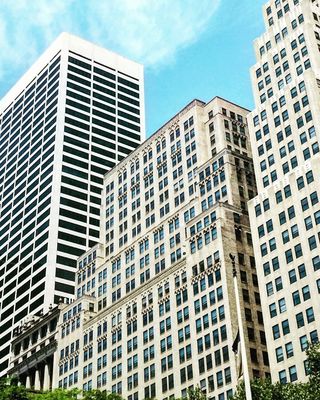 Tall city skyscrapers under a bright blue sky with scattered clouds, surrounded by green trees.