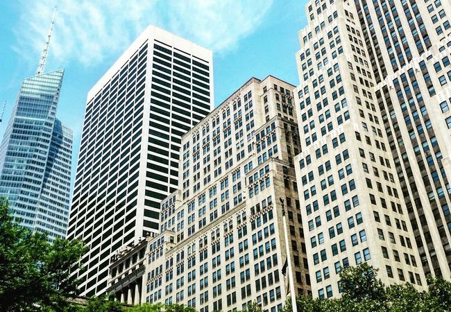Tall city skyscrapers under a bright blue sky with scattered clouds, surrounded by green trees.