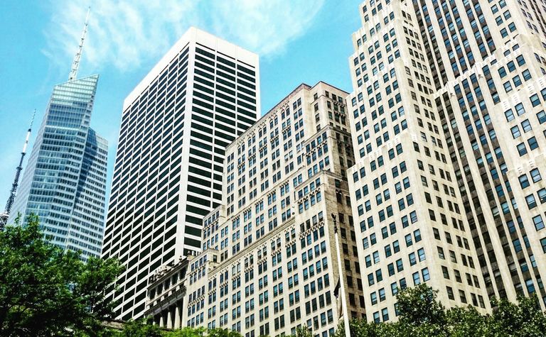 Tall city skyscrapers under a bright blue sky with scattered clouds, surrounded by green trees.
