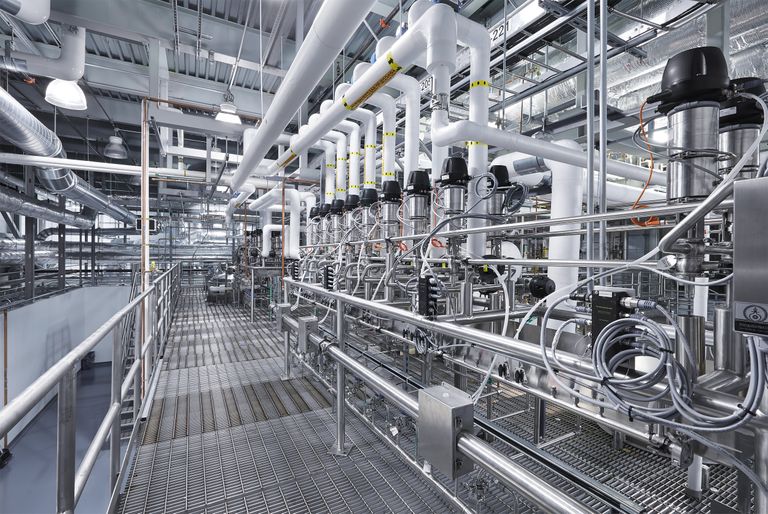 Interior of a modern industrial facility with metal walkways, pipelines, and machinery, featuring intricate piping and valves.