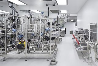 Industrial laboratory with stainless steel machinery, intricate piping, control panels, and red valves, under bright ceiling lights.
