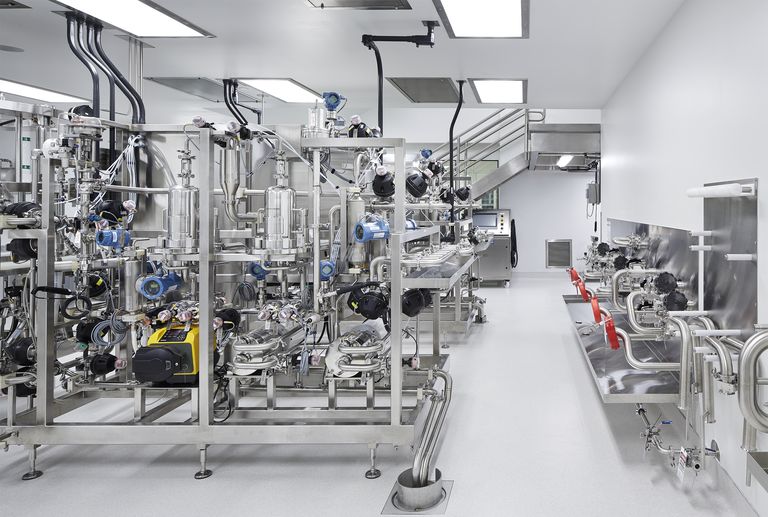 Industrial laboratory with stainless steel machinery, intricate piping, control panels, and red valves, under bright ceiling lights.