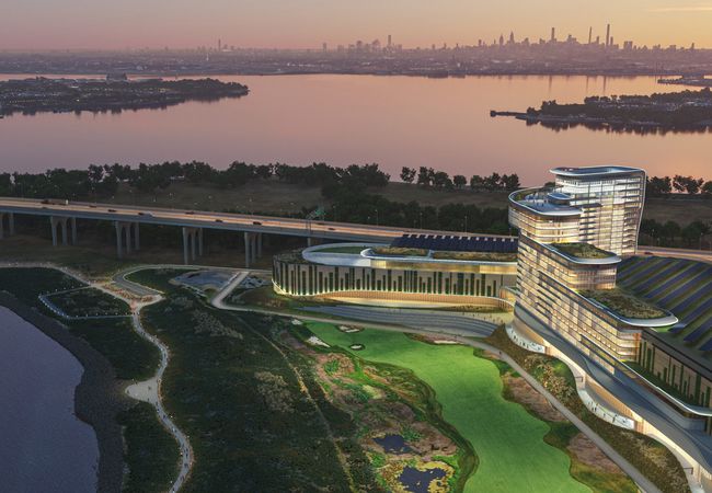 Modern waterfront building with curved architecture, surrounded by greenery and water, at dusk with a city skyline in the background.