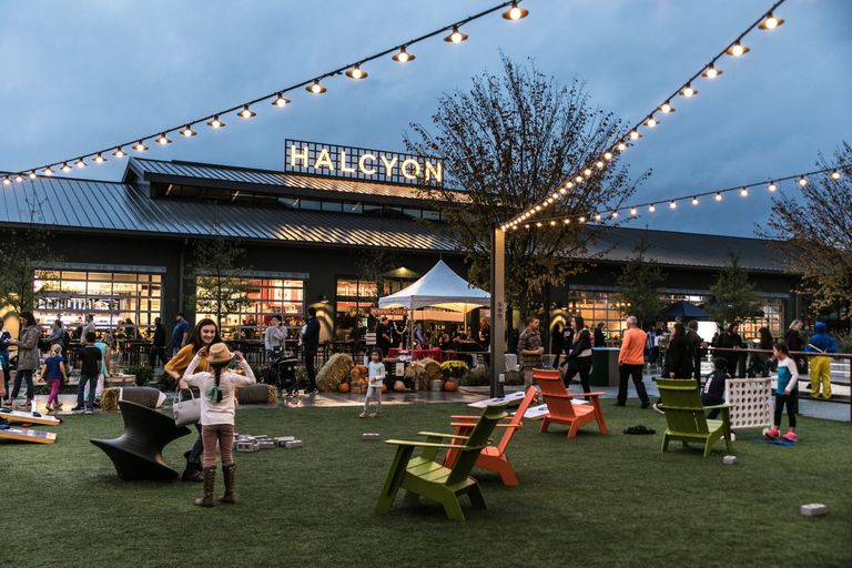 People gather in an outdoor area with string lights and colorful chairs, in front of a building with the sign "Halcyon" during twilight.