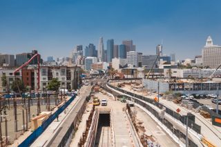 Construction site with cranes and vehicles in an urban area, with tall skyscrapers in the background under a clear blue sky.