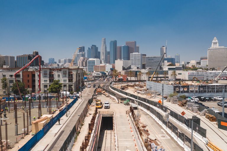 Construction site with cranes and vehicles in an urban area, with tall skyscrapers in the background under a clear blue sky.