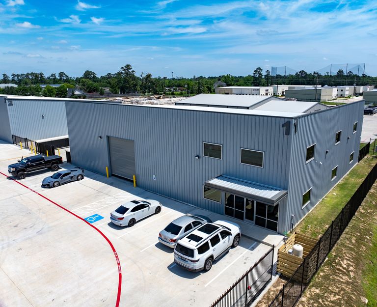 Industrial building with a parking lot, featuring several parked cars, clear blue sky, and a surrounding green area.