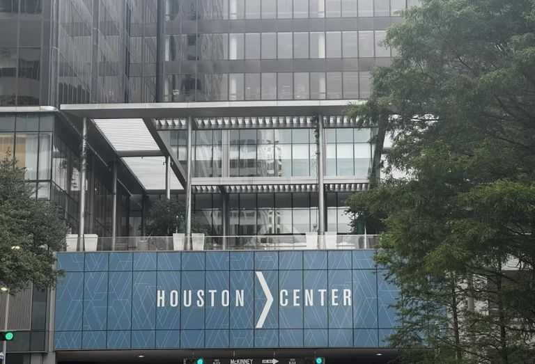 Exterior view of Houston Center, featuring a modern glass facade with a blue sign displaying the building's name, and trees in the foreground.