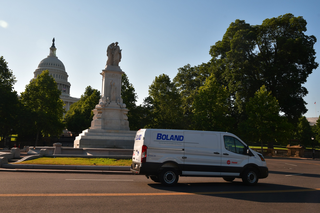 A white van drives past a statue in front of the U.S. Capitol building, surrounded by trees on a sunny day.