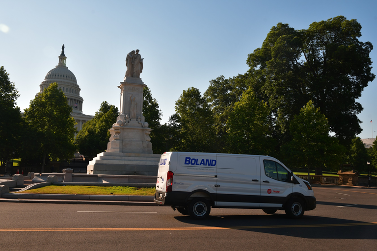 A white van drives past a statue in front of the U.S. Capitol building, surrounded by trees on a sunny day.