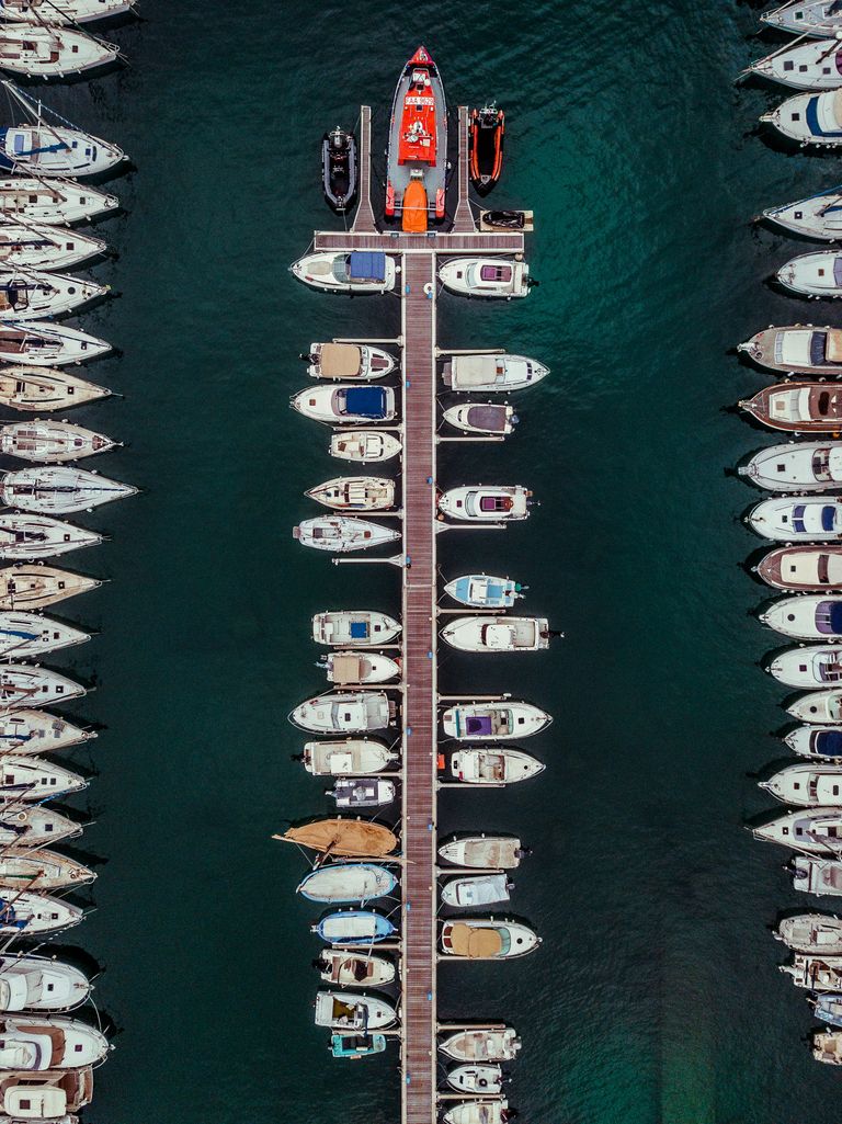 Aerial view of a marina with a central dock lined with various boats and a bright red boat near the end, surrounded by green water.
