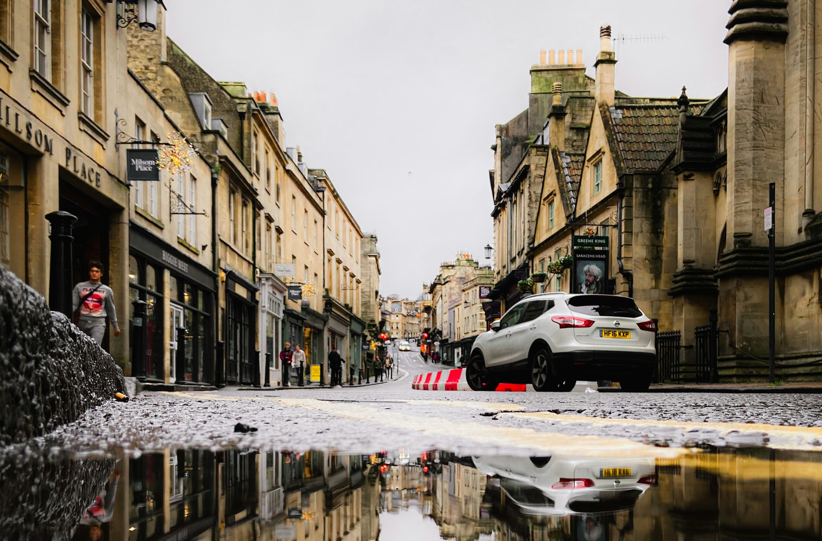 A flooded street in a historic town with buildings and a car reflects in a large puddle on a cloudy day.