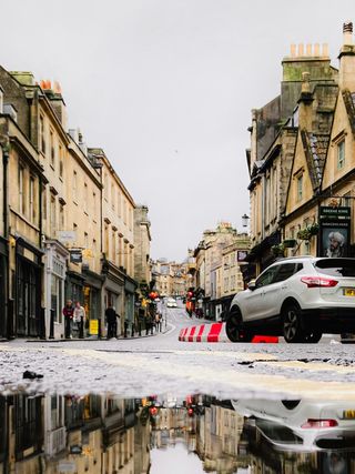 A flooded street in a historic town with buildings and a car reflects in a large puddle on a cloudy day.