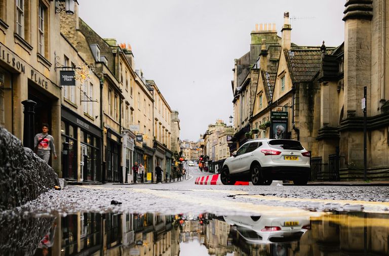 A flooded street in a historic town with buildings and a car reflects in a large puddle on a cloudy day.