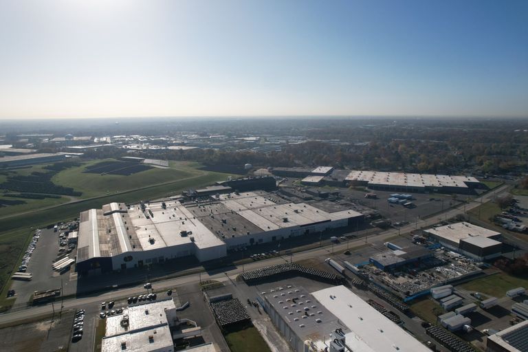 Aerial view of a large industrial complex with multiple warehouses, surrounded by green areas and a distant cityscape under a clear sky.