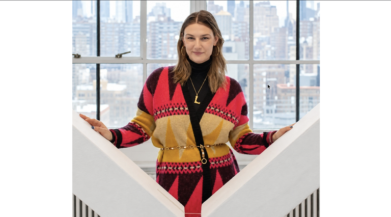 A person stands smiling in front of a window overlooking a cityscape, wearing a colorful geometric-patterned cardigan with a gold chain belt.