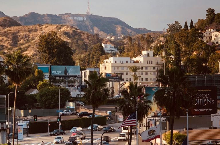 View of the Hollywood sign and palm trees, traffic, and billboards along Highland Avenue in the foreground, with the Hollywood Sign and hills in the background under a clear sky.