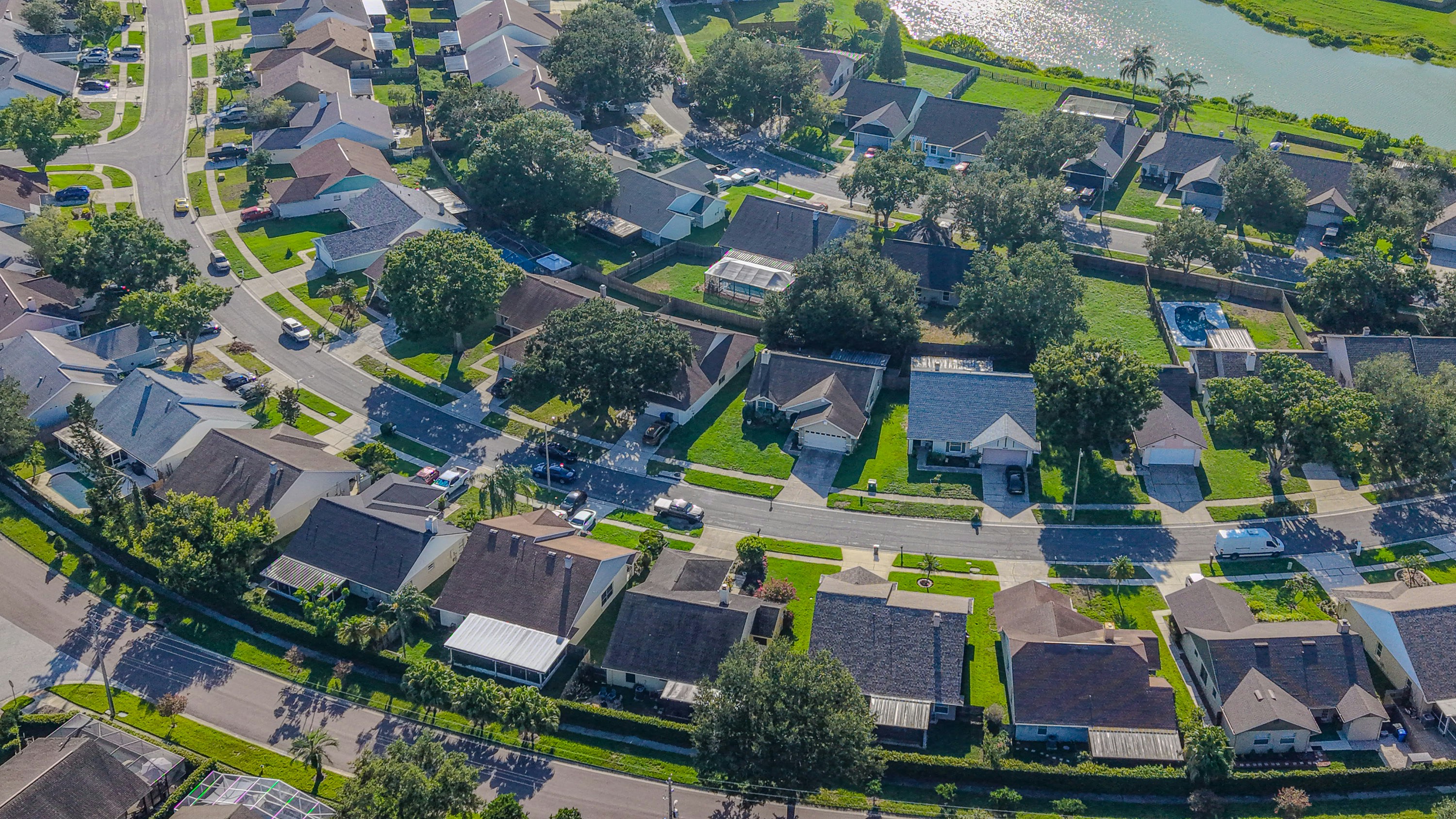 Aerial view of a suburban neighborhood with houses, roads and trees, adjacent to a body of water with green spaces.