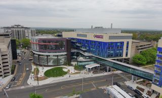Aerial view of a modern hospital complex with colorful facades, a curved glass walkway, and adjacent roads bordered by trees and other buildings.