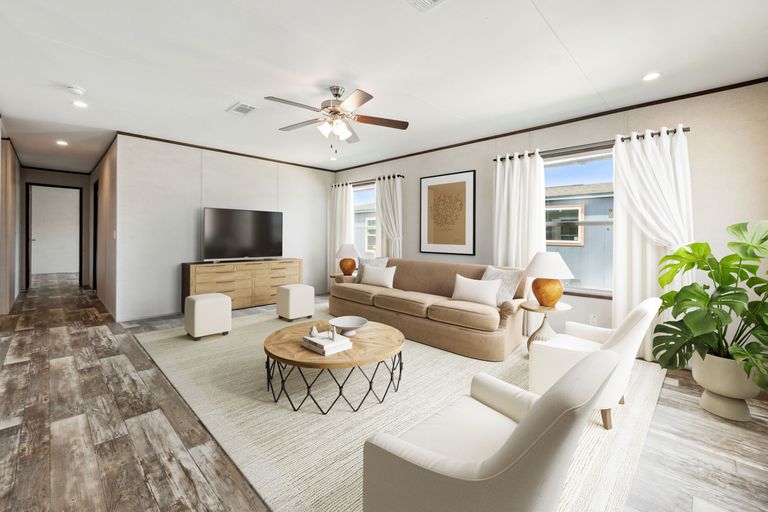 Living room with beige sofa, white armchairs, wooden coffee table, TV, large plant, and curtains. Ceiling fan and wooden flooring.