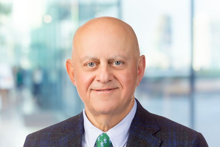 An older man in a suit and tie smiles at the camera, standing in a light, modern interior with blurred glass walls in the background.