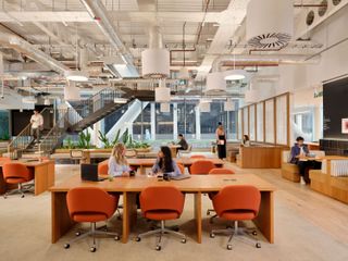 Modern office space with open seating, orange chairs, large windows, plants, and employees working and walking around.
