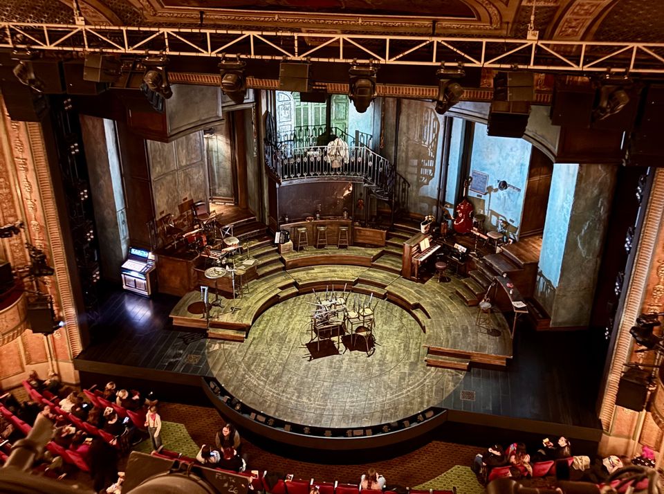 View of a theater stage set with musical instruments, a piano, and chairs arranged in a semi-circle, under overhead lights. Audience seated below.