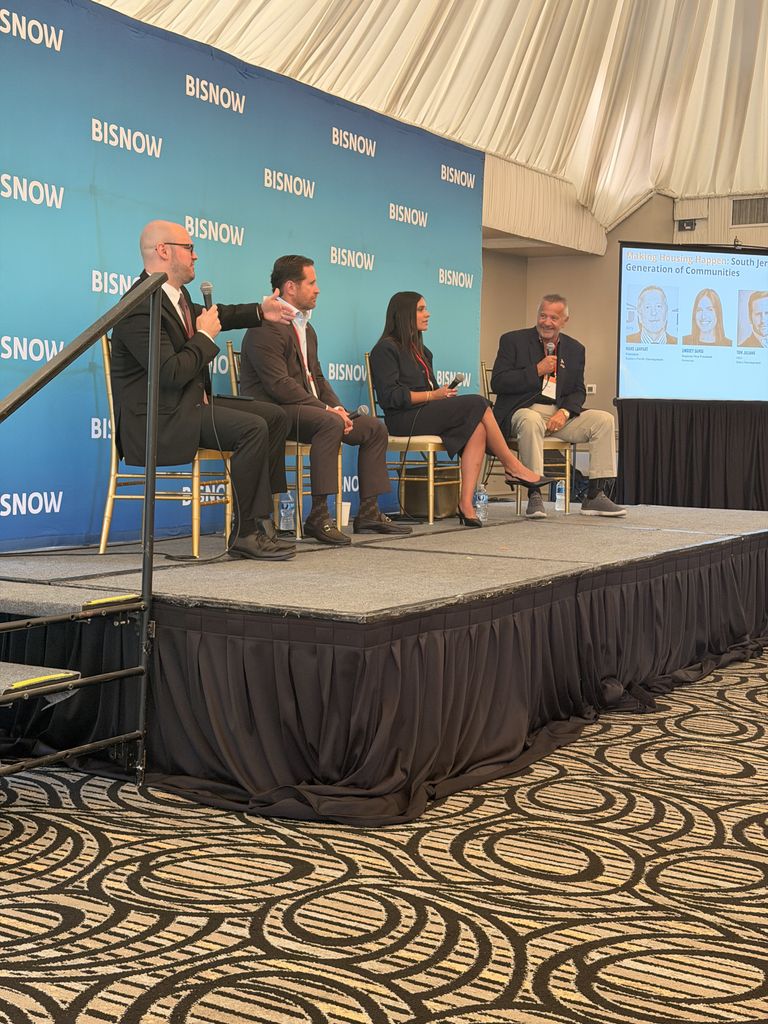 A panel of four professionals sits on a stage discussing "Making Housing Happen" at a Bisnow event, with a display screen showing speaker details.