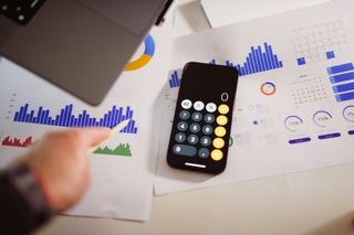 A smartphone with a calculator app lies on a desk next to printed graphs and charts, while a hand with a pen points at a bar graph.