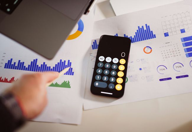 A smartphone with a calculator app lies on a desk next to printed graphs and charts, while a hand with a pen points at a bar graph.