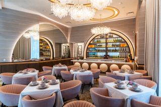 Elegant restaurant with pink velvet chairs, white tablecloths, chandeliers, and a backlit bar displaying wine bottles against mirrored walls.