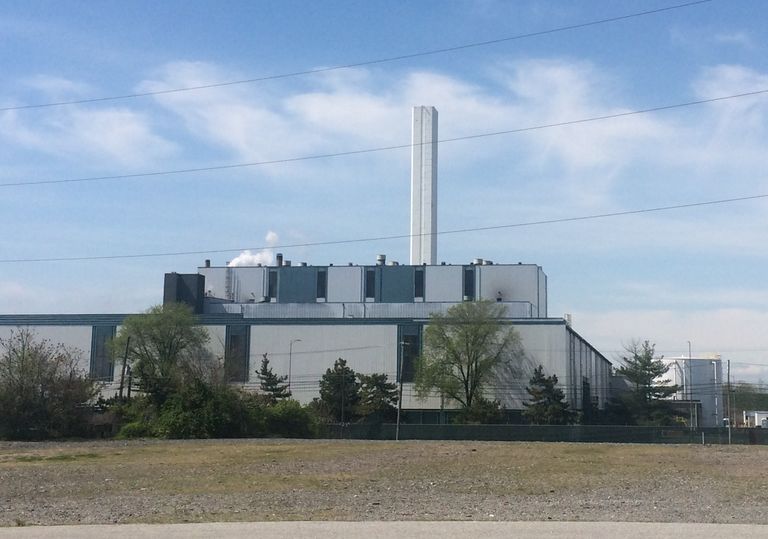 Industrial building with a tall chimney emitting smoke, surrounded by trees under a clear blue sky with wispy clouds.