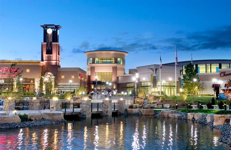 Night view of a shopping center with illuminated signs and a clock tower. Reflections on a pond in the foreground enhance the scene.