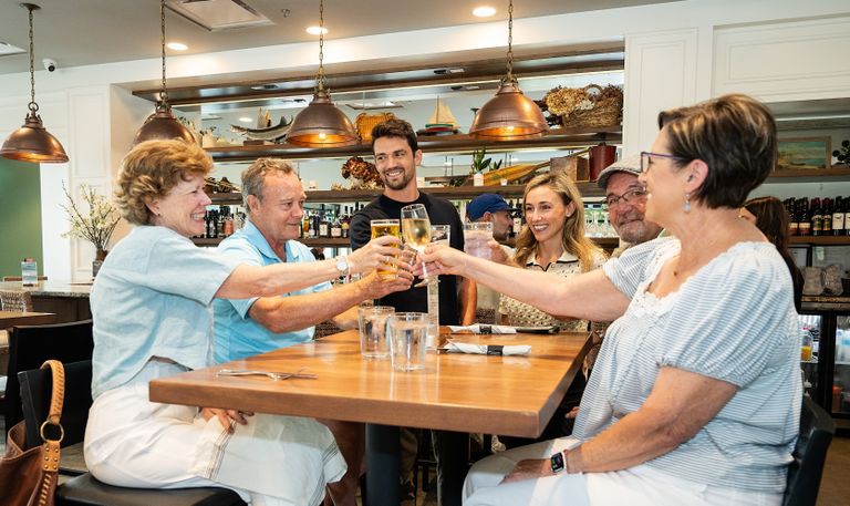 A group of six people clink glasses in a cheerful toast while seated at a wooden table in a cozy, well-lit restaurant.