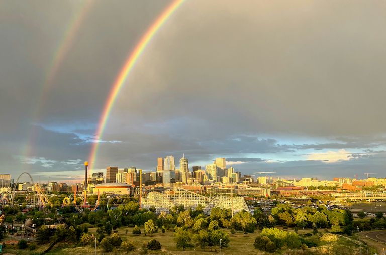 A vibrant double rainbow arcs over a city skyline at sunset, with scattered clouds and amusement park rides visible in the foreground.
