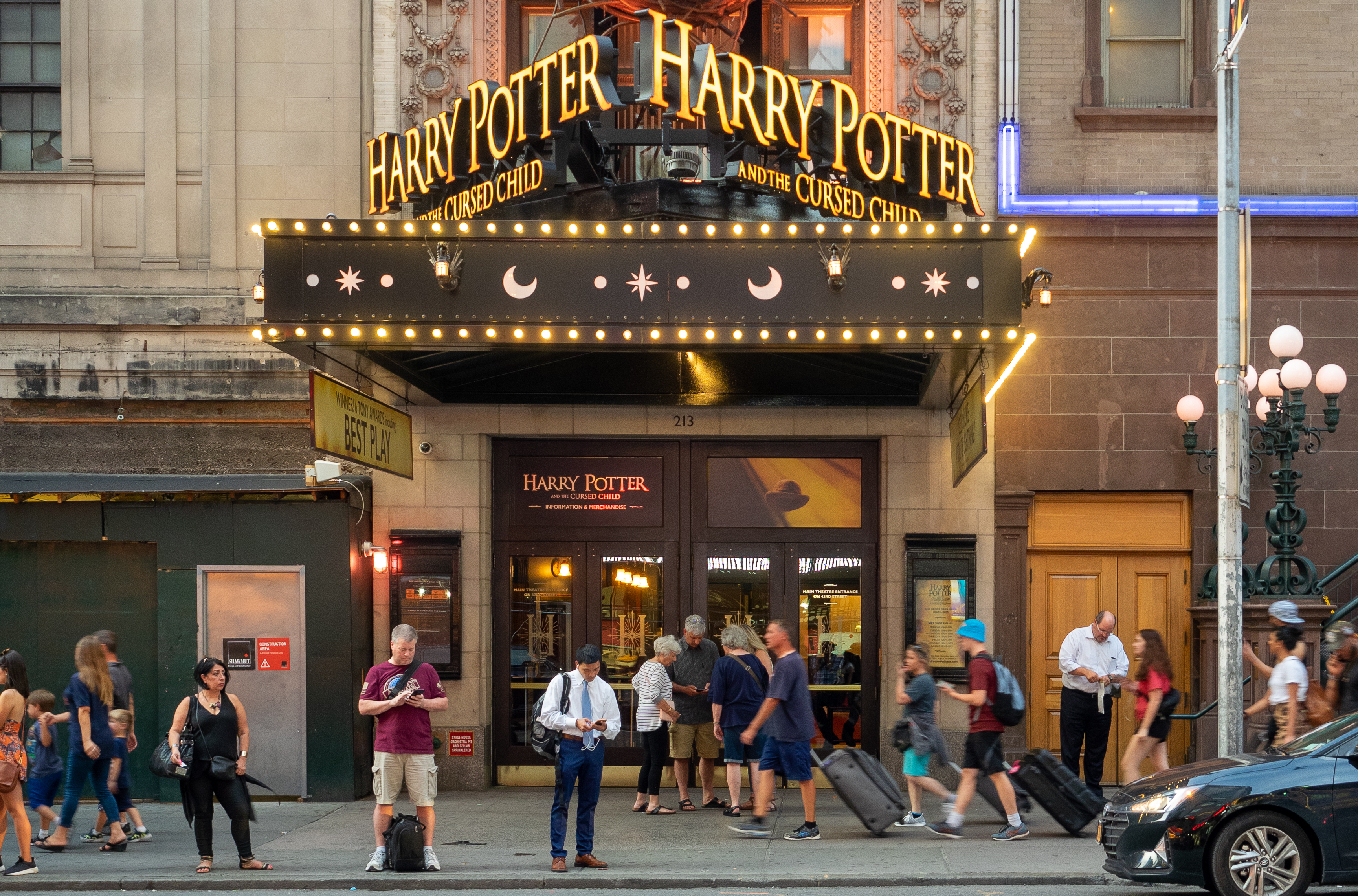 People walk past a theater entrance with "Harry Potter and the Cursed Child" signage, featuring decorative lights and a busy urban scene.