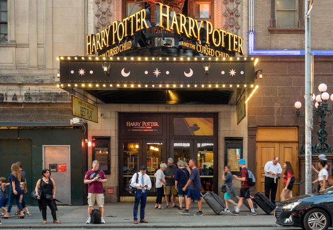 People walk past a theater entrance with "Harry Potter and the Cursed Child" signage, featuring decorative lights and a busy urban scene.