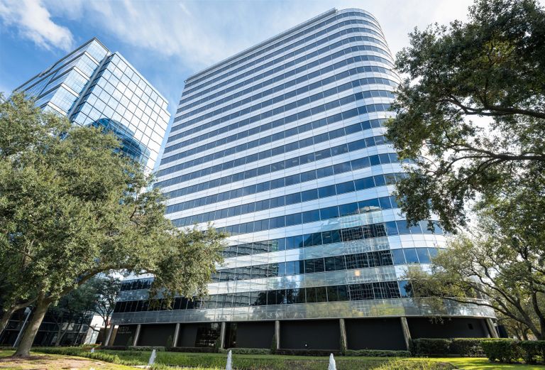 Tall, modern glass office building with rounded corners against a blue sky, surrounded by green trees and shrubs.
