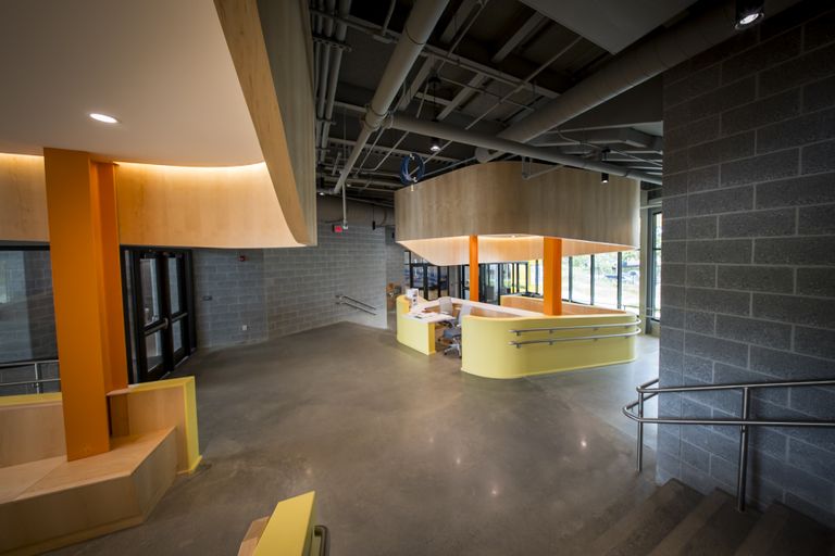 Modern lobby with orange and yellow accents, featuring a central reception desk, concrete floors, and large windows.