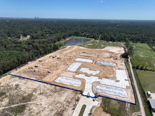 Aerial view of a construction site with foundations laid, surrounded by a forested area and a pond, under a clear blue sky.