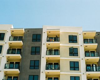 Modern apartment building with balconies, featuring a person with colorful clothing standing on one of the upper balconies under a blue sky.