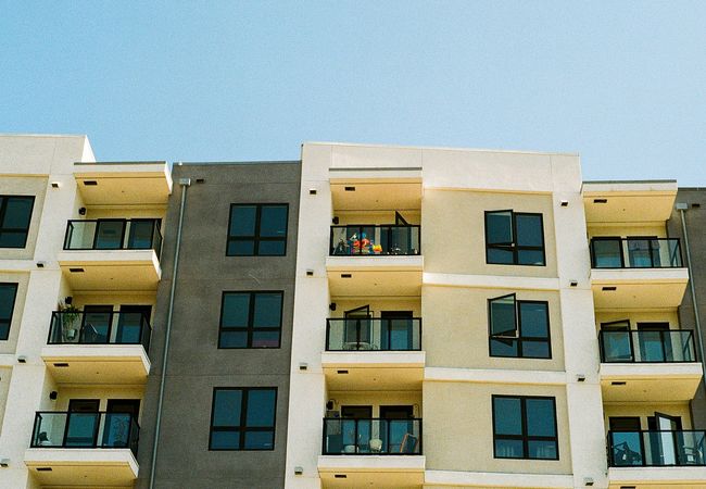 Modern apartment building with balconies, featuring a person with colorful clothing standing on one of the upper balconies under a blue sky.