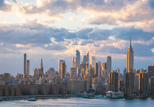 Skyline of a city at sunset with skyscrapers reflecting golden light, under a partly cloudy sky and waterfront in the foreground.