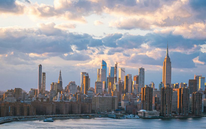 Skyline of a city at sunset with skyscrapers reflecting golden light, under a partly cloudy sky and waterfront in the foreground.