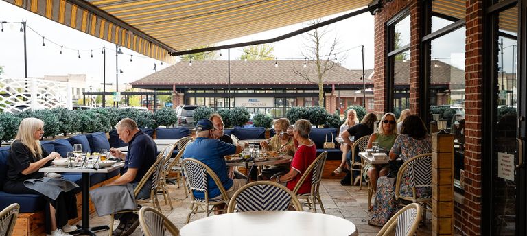 People dining at outdoor restaurant tables under a striped awning, with plants and string lights creating a cozy atmosphere.
