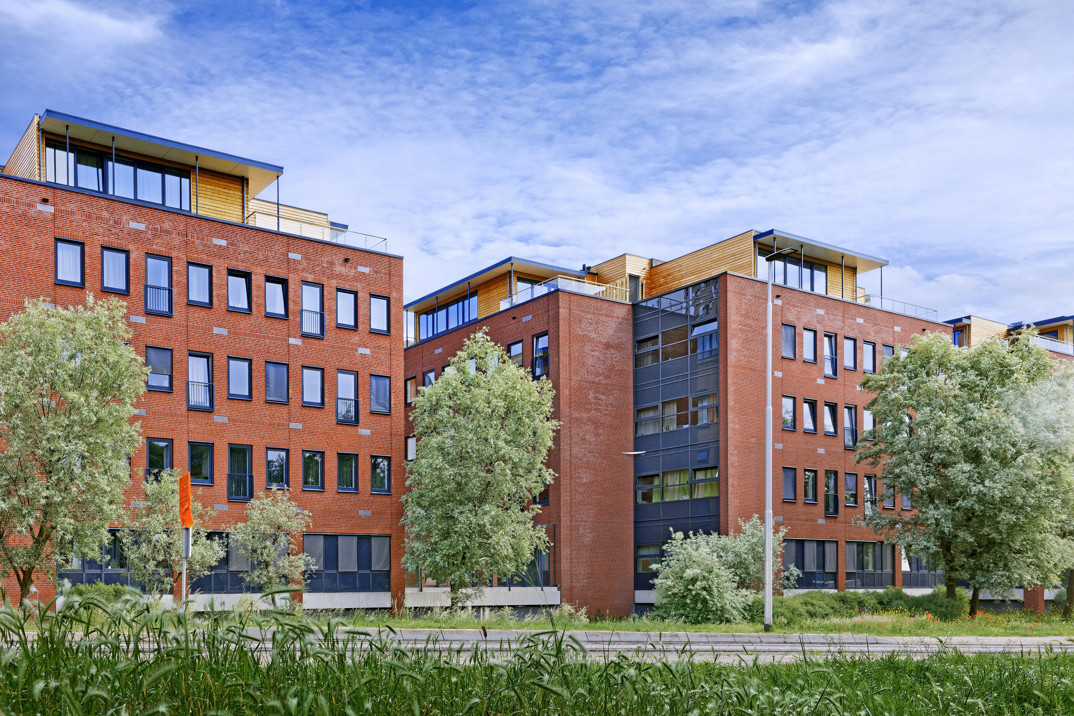 A modern red brick office building with multiple floors and large windows, surrounded by trees and greenery, set against a partly cloudy sky.
