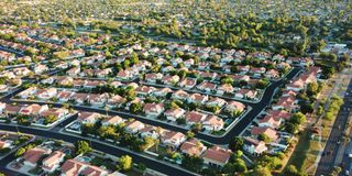 Aerial view of a suburban neighborhood with uniform red-roofed houses, lush green trees, and neatly arranged streets extending into the distance.
