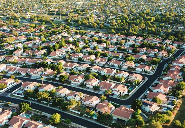 Aerial view of a suburban neighborhood with uniform red-roofed houses, lush green trees, and neatly arranged streets extending into the distance.