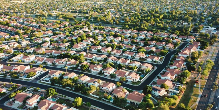 Aerial view of a suburban neighborhood with uniform red-roofed houses, lush green trees, and neatly arranged streets extending into the distance.