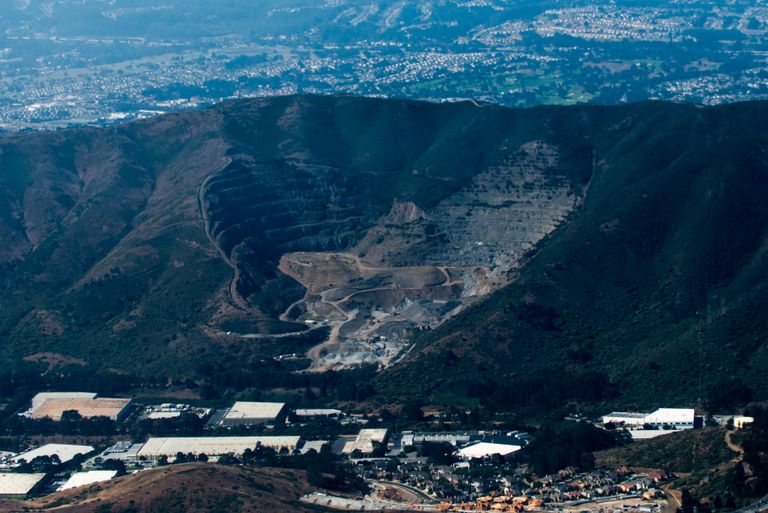 Aerial view of a large quarry in a mountainous area surrounded by urban development and green hills under a clear sky.